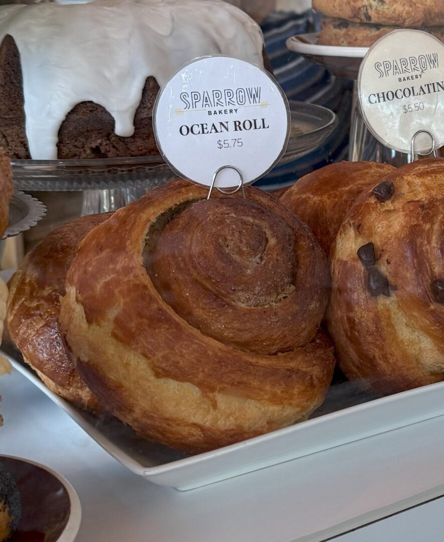 Sparrow Bakery Ocean Roll a Close Up of a Golden Brown Pastry Labeled Ocean Roll 5 from Sparrow Bakery Displayed on a White Tray the Pastry is Swirled and Flaky Sitting Beside Other Baked Goods Including a Chocolate Pastry and a Cake with White Icing