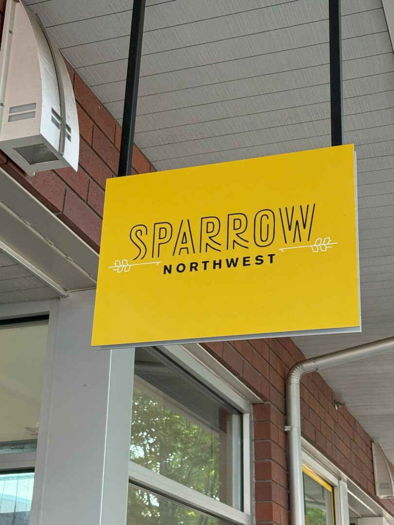 a Yellow Hanging Sign with sparrow Northwest in Bold Black Letters and Simple Decorative Lines Mounted Under a Covered Walkway with a Red Brick Wall and Windows in the Background