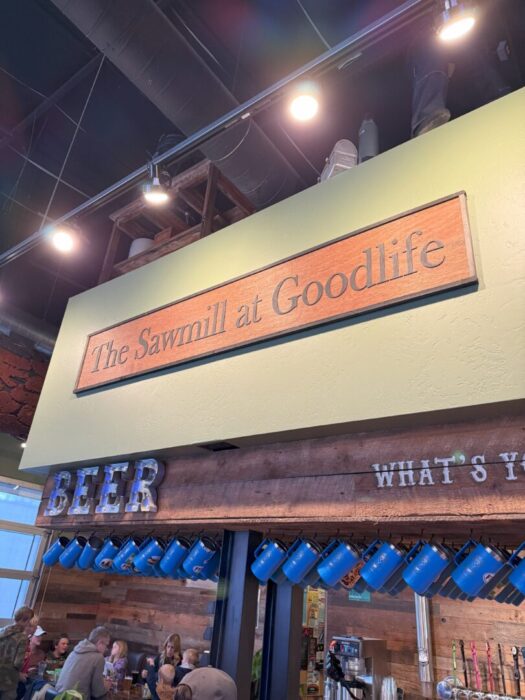 a Wooden Sign Reading the Sawmill at Goodlife is Mounted Above a Bar Area Below a Rustic Beer Sign and Blue Mugs Hang While People Are Gathered and Seated at Tables the Ceiling and Lights Are Visible Above the Green Wall a Wooden Sign Reading the Sawmill at Goodlife is Mounted Above a Bar Area Below a Rustic Beer Sign and Blue Mugs Hang While People Are Gathered and Seated at Tables the Ceiling and Lights Are Visible Above the Green Wall
