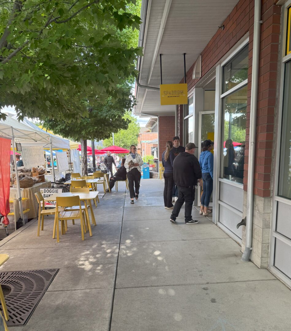 Waiting Line Sparrow Bakery People Stand in Line Outside a Storefront with a Yellow Sign Yellow Chairs and Tables Are on the Sidewalk Under a Tree to the Left Vendors with White Tents and Red Umbrellas Are Set Up at a Market Its a Sunny Day with Green Trees Along the Walkway