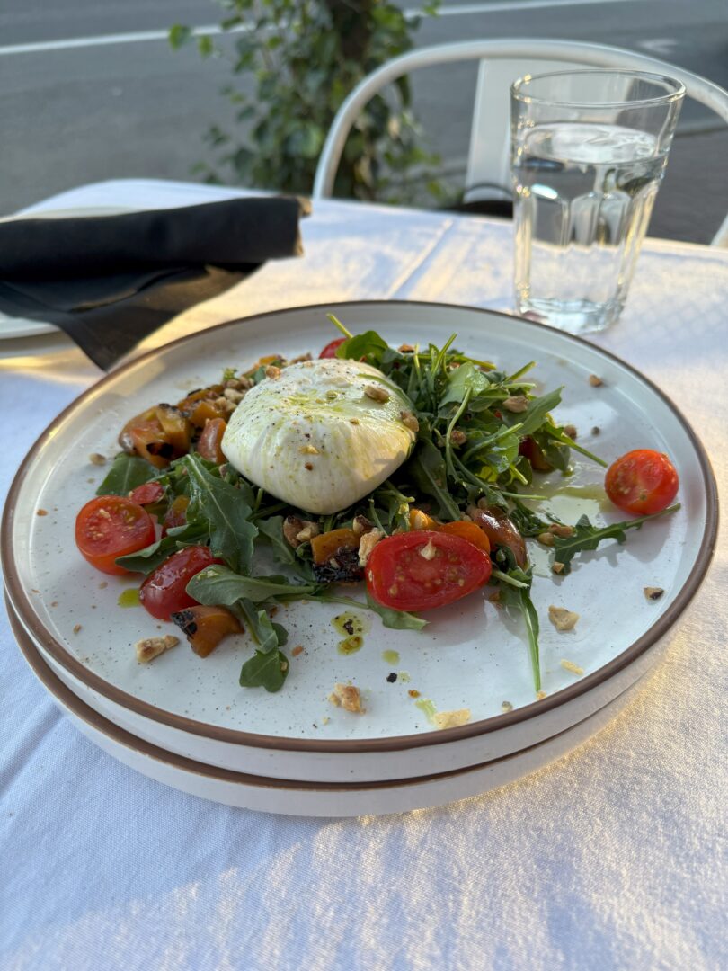 A plate of salad with burrata cheese on top, surrounded by arugula, cherry tomatoes, nuts, and roasted vegetables. Enjoy this fresh dish outdoors at one of the top Bend Restaurants, with a glass of water and a fork wrapped in a black napkin.