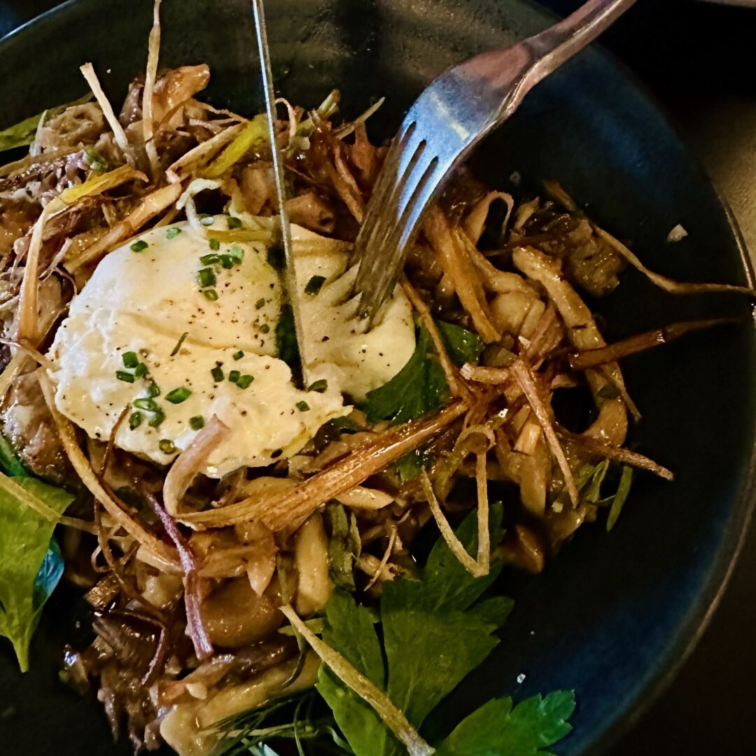 A dark bowl filled with sautéed mushrooms, crispy fried thin root vegetables, fresh herbs, and topped with a runny poached egg, which is being pierced by a fork. The dish has an earthy, rustic presentation with vibrant greens.