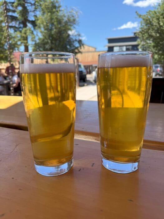 Two Tall Glasses of Golden Beer with Foamy Heads Sit on a Wooden Outdoor Table Sunlight Streams Through Reflecting the Vibrant Scene in the Background People Enjoy a Sunny Daya Perfect Setting for Sampling Brews from Bend Breweries