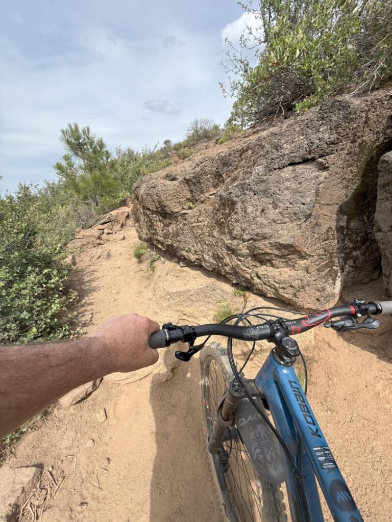 A person rides a blue mountain bike on the narrow, rocky Rimrock Trail. Their left hand grips the handlebar as rugged rocks and green shrubs line the path under a partly cloudy sky, evoking outdoor adventure and mountain biking.