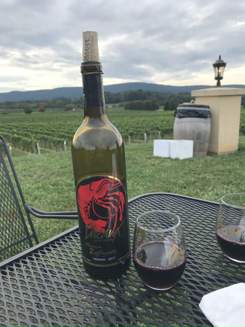 Breaux vineyards loudoun county a Bottle of Red Wine Labeled with a Red Scorpion Graphic Stands on a Metal Patio Table at Market Table Bistro Alongside Two Glasses of Red Wine Behind a Vineyard Stretches out Under Cloudy Skies with Green Hills in the Distance and a Barrel and Lantern Nearby