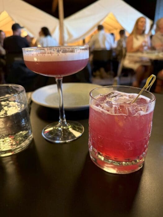 Two pink cocktails sit on a dark table: one is in a coupe glass with foam on top, the other in a short glass with ice and a cocktail pick. A glass of water, an empty plate, and people dining inside a warmly-lit tented space are visible in the background.