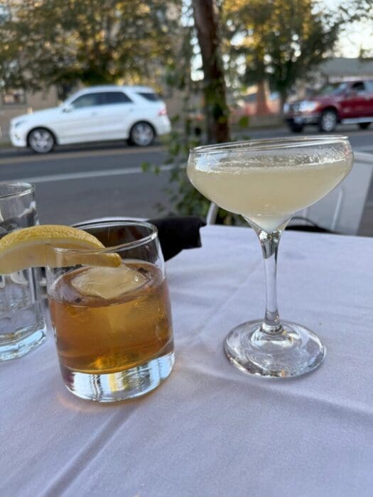 Two Cocktails Sit on a White Tablecloth Outdoorsan Old Fashioned with a Lemon Twist and a Pale Yellow Drink in a Coupe Its the Perfect Scene for Summer Evenings at Some of the Best Bars in Bend Or with Cars and Trees Lining the Street Behind