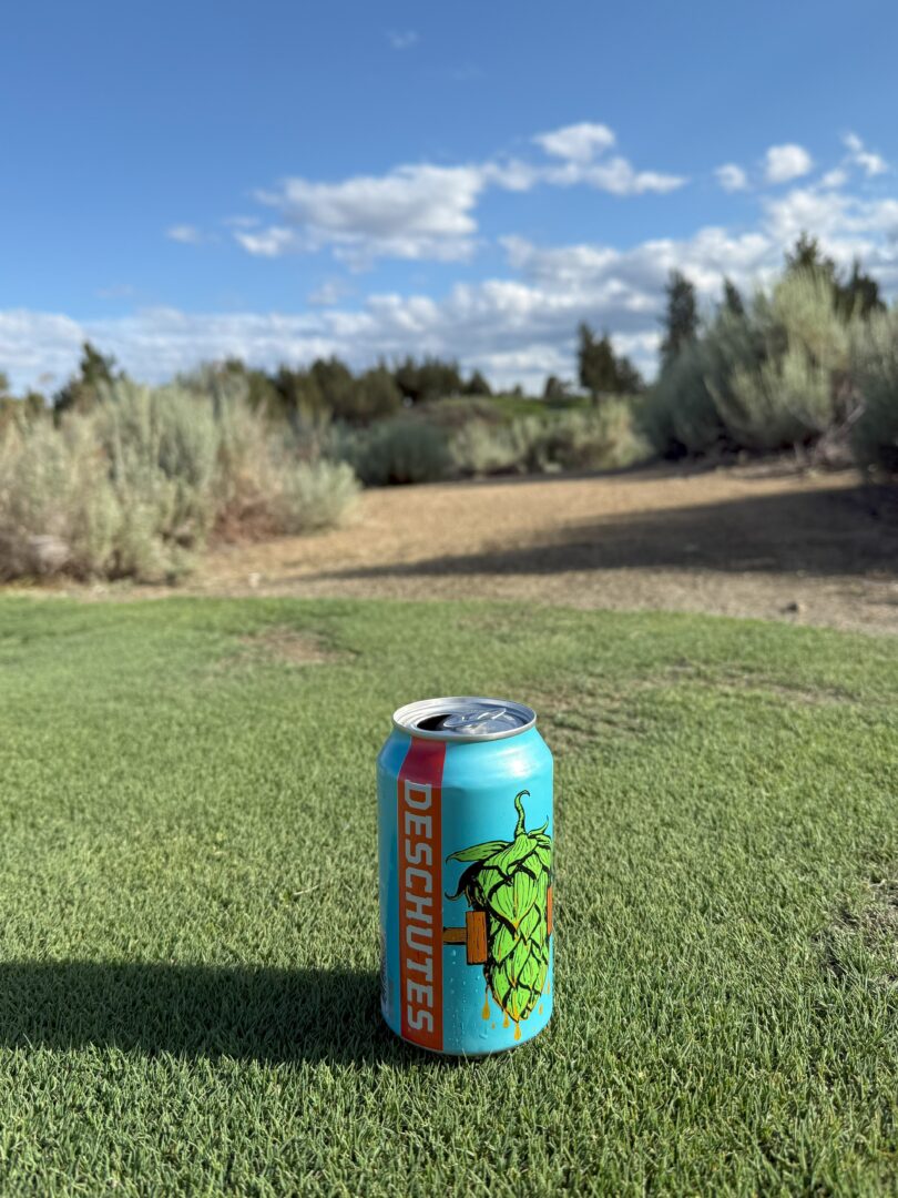 Deschutes Beer Pronghorn Golf Course a Blue Deschutes Beer Can with a Green Hop Illustration Stands on Green Grass in the Foreground Reminiscent of a Bend Oregon Golf Course Behind It Shrubs and Dirt Appear Beneath a Bright Blue Sky with Scattered White Clouds