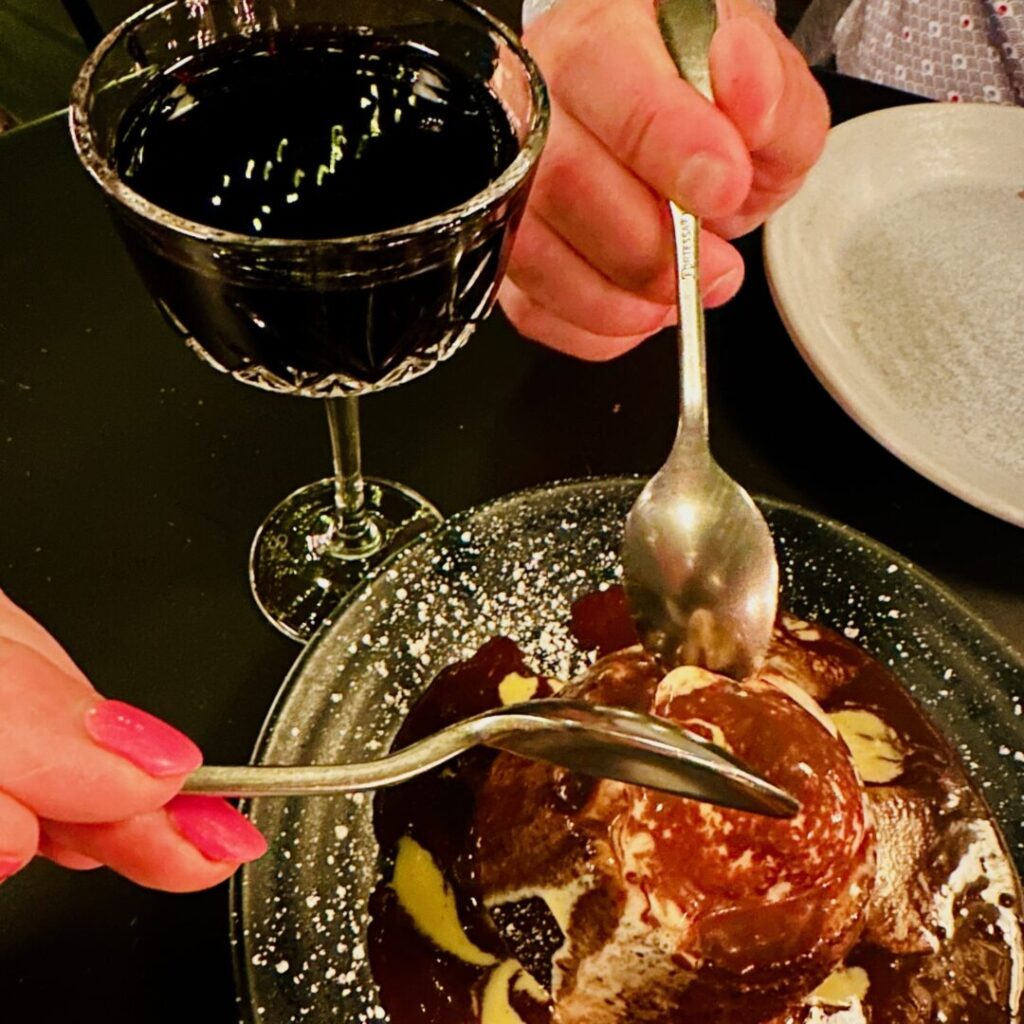 A close-up of two people sharing a chocolate dessert with spoons, next to a crystal glass of red wine on a dark table. The dessert is topped with chocolate sauce and powdered sugar. One person has pink nails; both plates and hands are partially visible.