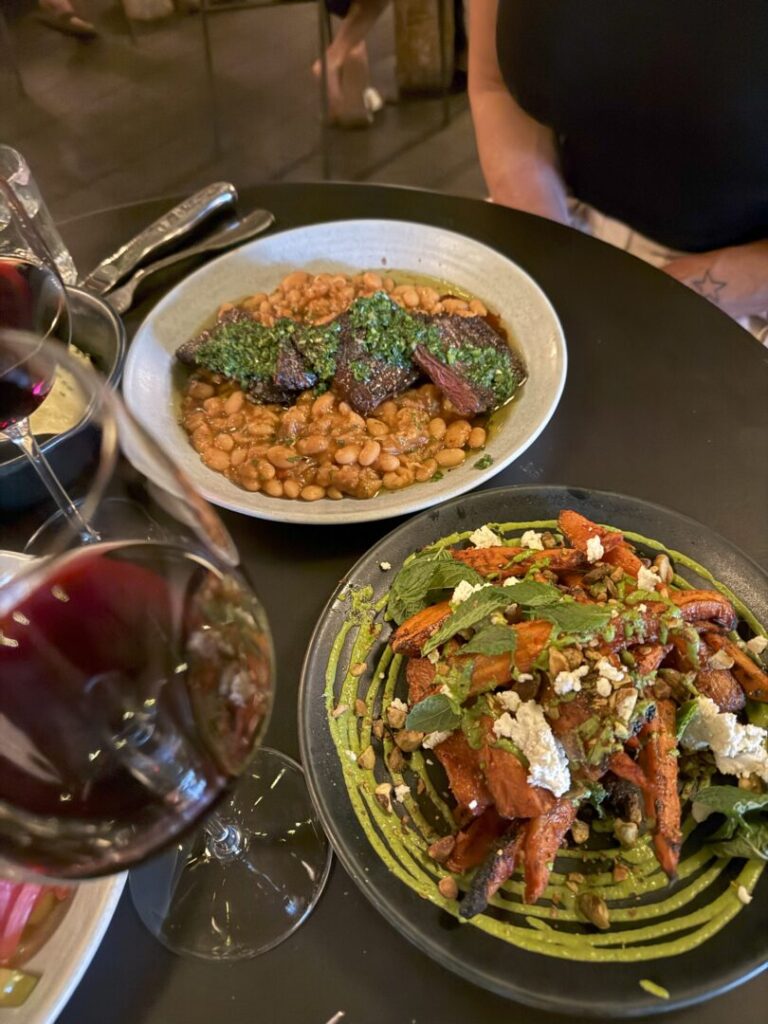 Two glasses of red wine on a dark table beside two dishes: one with braised meat and beans in a green herb sauce, and another with roasted carrots topped with greens, nuts, and crumbled cheese. A person in a sleeveless top sits in the background.