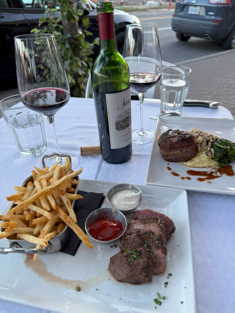 A table set for two outdoors at one of the top Bend Restaurants, featuring a bottle of red wine, fries with ketchup, sliced steak, and a dish with filet mignon, mashed potatoes, mushrooms, and broccolini.