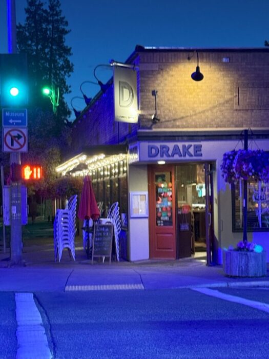 a Cozy Restaurant Called Drake Sits on a Street Corner at Dusk in Bend Warm String Lights Above Its Patio Evoke the Inviting Charm Typical of Bend Restaurants Stacked White Chairs a Red Umbrella and a Glowing Green Traffic Light Complete the Scene