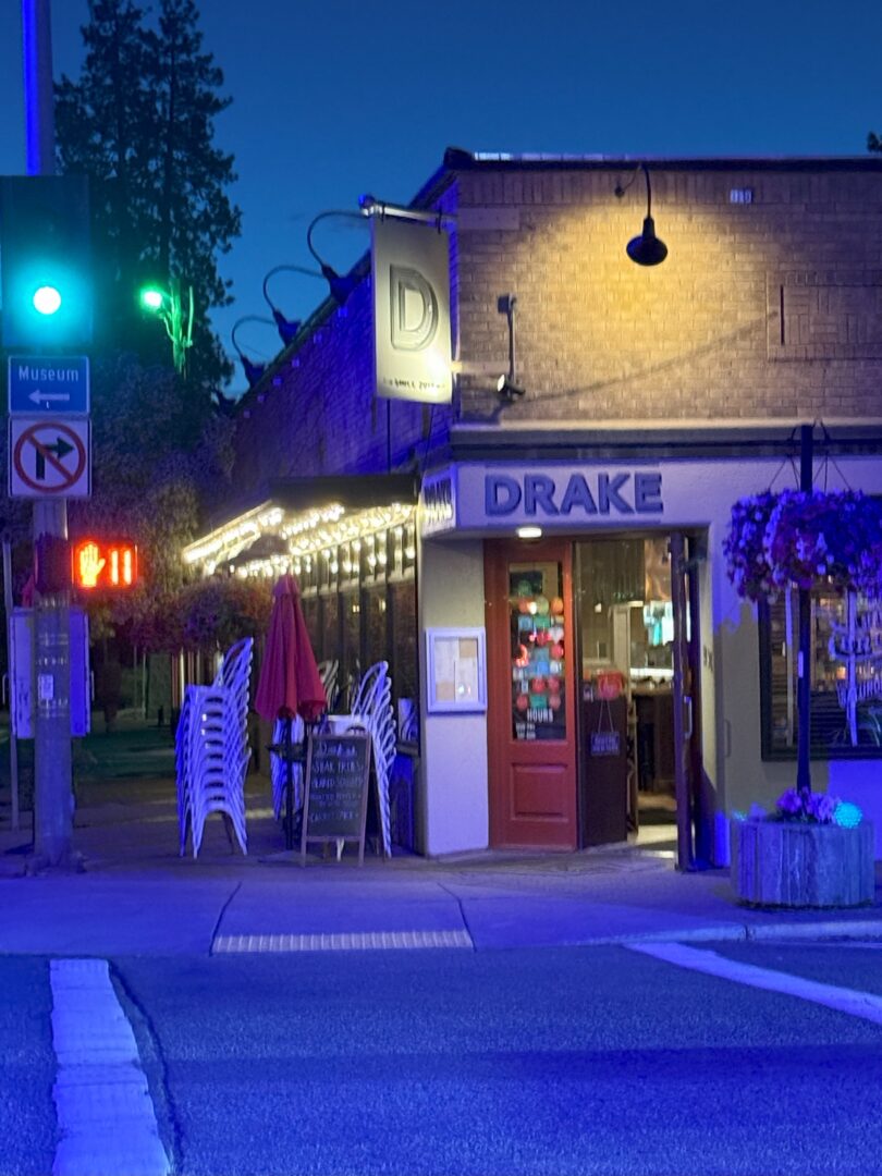 A cozy restaurant called DRAKE sits on a street corner at dusk in Bend. Warm string lights above its patio evoke the inviting charm typical of Bend restaurants. Stacked white chairs, a red umbrella, and a glowing green traffic light complete the scene.