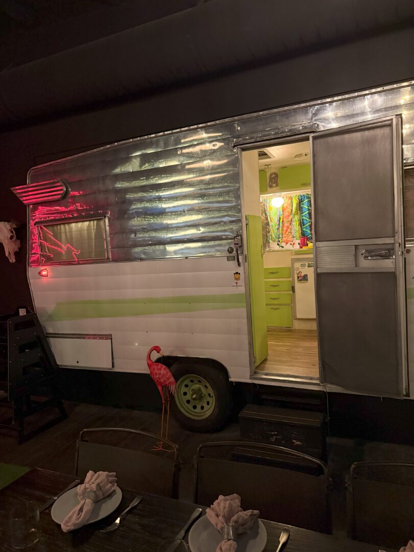 A shiny silver and white vintage camper with a green stripe is parked indoors. Its door is open, revealing a brightly lit interior with colorful decor. A pink flamingo lawn ornament stands outside. A dining table with napkins folded on plates is in the foreground.