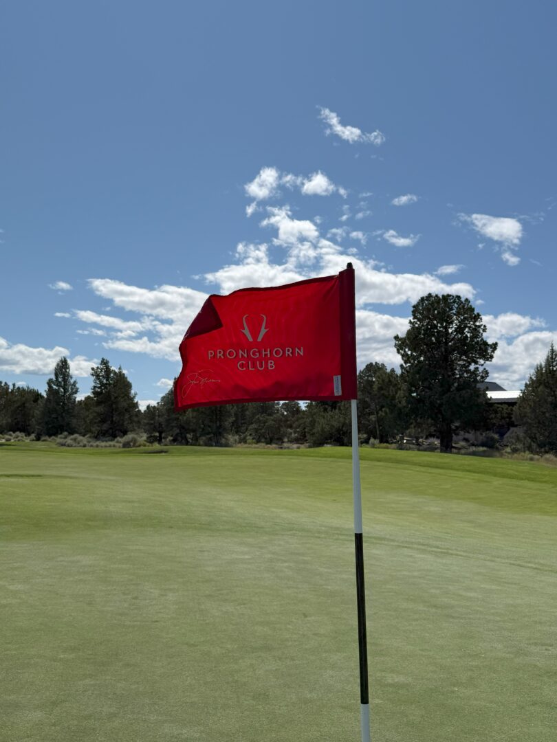 Flag Stick Pronghorn Golf Club a Red Flag Reading Pronghorn Club Flutters Atop a Flagstick on the Green at Pronghorn Golf Course the Mostly Clear Sky and Tree Lined Background Are Bathed in Bright Sunlight Casting Sharp Shadows Across the Well Manicured Grass