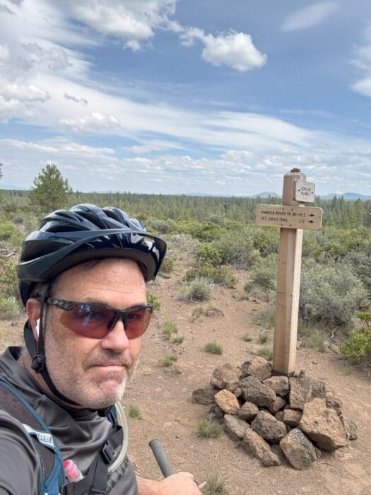 A man wearing a bike helmet, sunglasses, and a hydration pack stands in front of a Rimrock Trail sign and a pile of rocks on a dirt path, with shrubs and pine trees behind him under a partly cloudy sky.