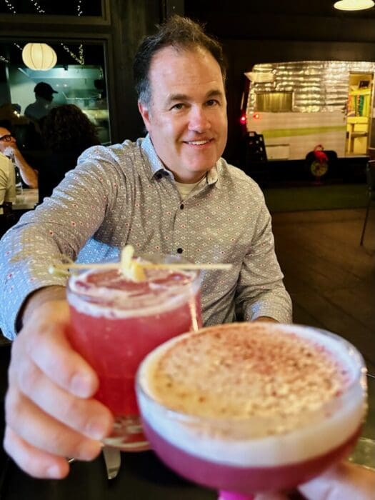 A smiling man in a patterned shirt sits at a table, holding up a pink cocktail with a garnish. Another hand in the foreground holds a similar pink drink, capturing the lively atmosphere of Bend restaurants, with string lights and a shiny food truck behind.