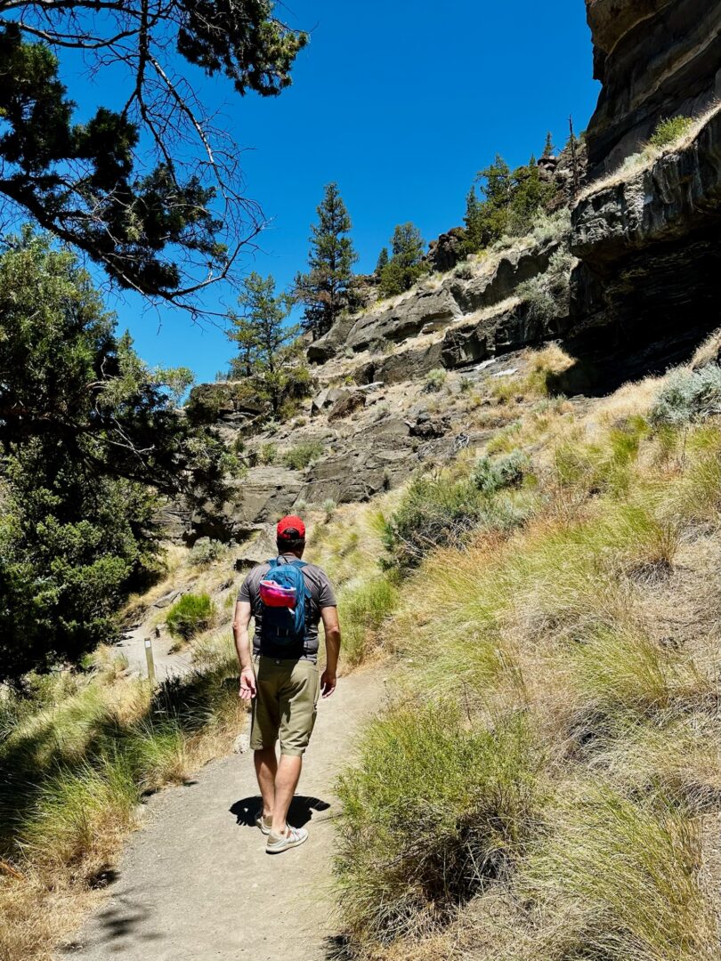 A person in a red cap, blue shirt, khaki shorts, and a backpack hikes up a sunlit dirt trail through dry grass and shrubs near Steelhead Falls, beside rugged cliffs and pine trees under a clear blue sky.