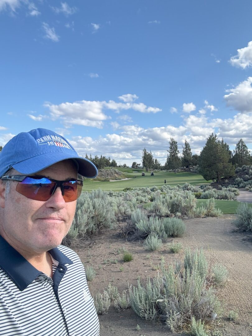 Glenn Pronghorn Golf Course a Man in a Blue Cap Sunglasses and Striped Polo Shirt Stands in the Foreground of Pronghorn Golf Course with Green Grass Sandy Patches Low Shrubs and Distant Trees Under a Bright Blue Sky with Scattered White Clouds