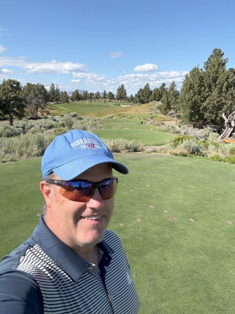 a Man Wearing a Blue Cap Sunglasses and a Striped Polo Shirt Smiles for a Selfie at Pronghorn Golf Course Behind Him Are Green Fairways Scattered Shrubs Pine Trees and a Bright Blue Sky with Some Clouds