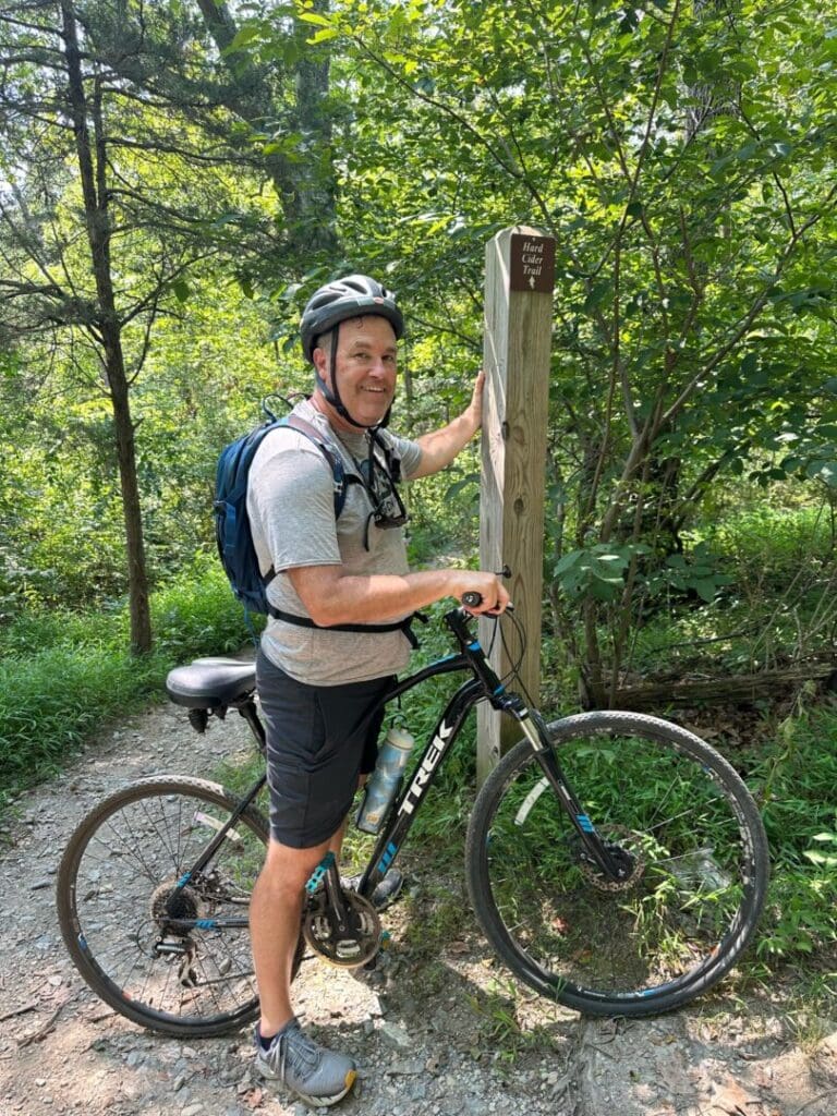 Glenn Vanlandingham Biking Little Bennet Maryland a Man in a Helmet and Backpack Stands Beside a Trek Mountain Bike on a Dirt Trail in the Mt Airy Md Forest He Smiles at the Camera While Holding a Wooden Trail Marker As Sunlight Highlights the Green Foliage Around Him