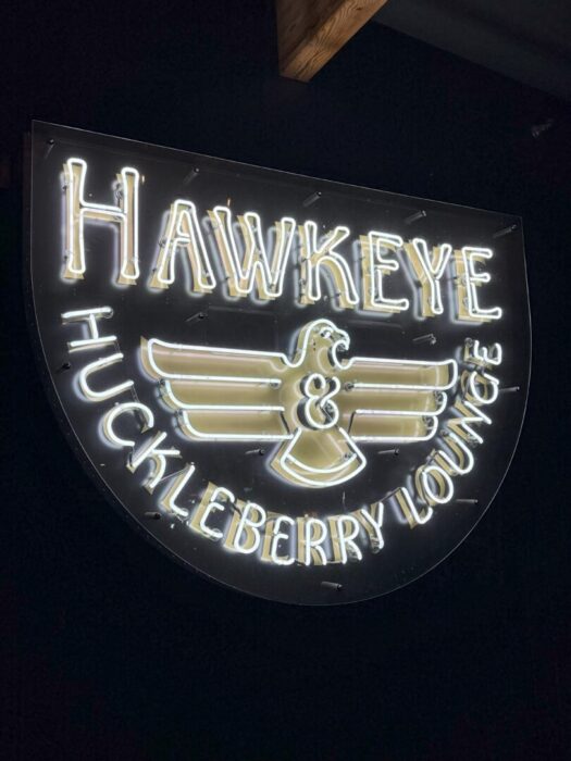 A glowing neon sign reads HAWKEYE & HUCKLEBERRY LOUNGE in white and gold, with a stylized bird spreading its wings. Set against a black background, this shield-shaped sign shines among Bend restaurants, outlined in radiant white neon.