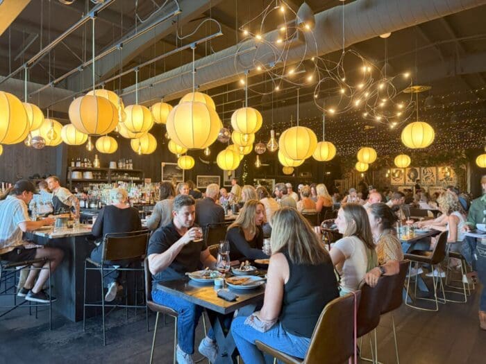 a Lively Restaurant Interior Reminiscent of Popular Bars in Bend Or with Many Diners at Tables and Bar Warmly Lit by Hanging Round Yellow Lanterns People Are Eating Drinking and Socializing Under Modern Decor and Twinkling String Lights