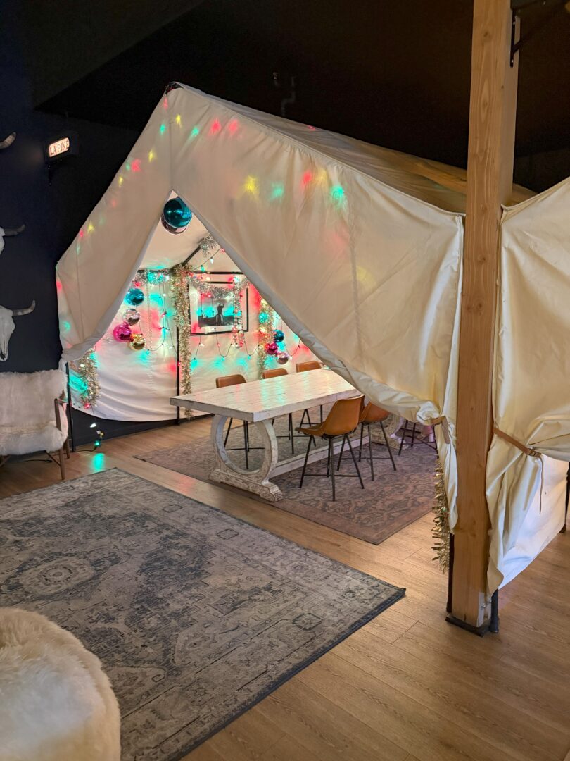 A cozy indoor tent with white fabric is decorated with colorful string lights and ornaments. Inside, a long table with mismatched chairs sits on a rug. The space has wood floors, a carpet, and soft ambient lighting, creating a festive, inviting atmosphere.
