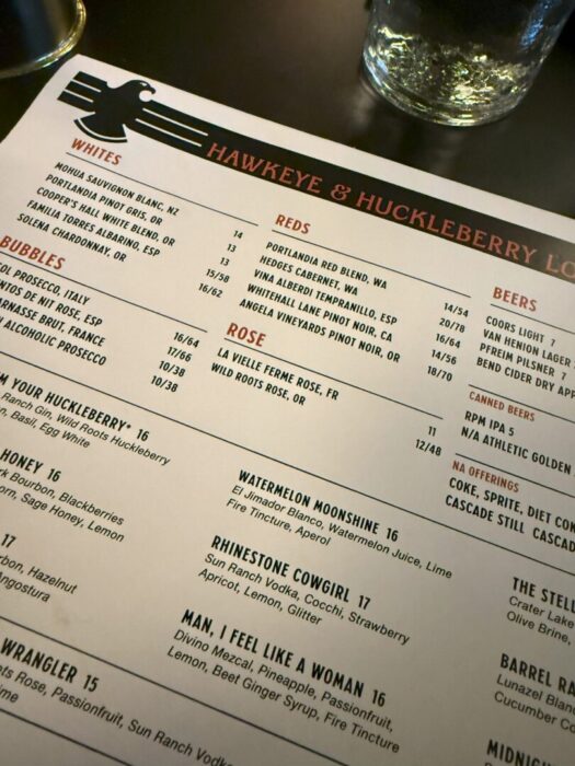 A close-up of a Hawkeye & Huckleberry Lounge menu on a dark table. Sections for wines, beer, and cocktails are visible, with a large black eagle graphic at the top. A glass of water sits at the upper right corner, partially visible.