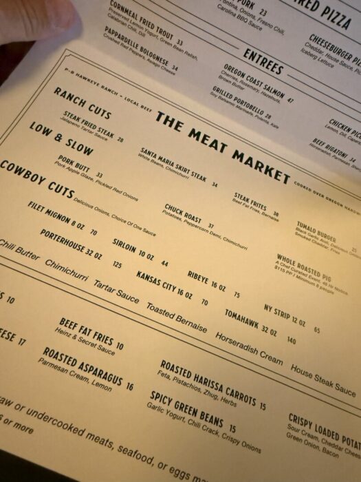 A close-up of a restaurant menu titled The Meat Market lists various steak and meat options, including Ranch Cuts, Cowboy Cuts, Low & Slow, and Steak Frites. Each item has a description and price. The menu is viewed at an angle, with yellowish lighting.