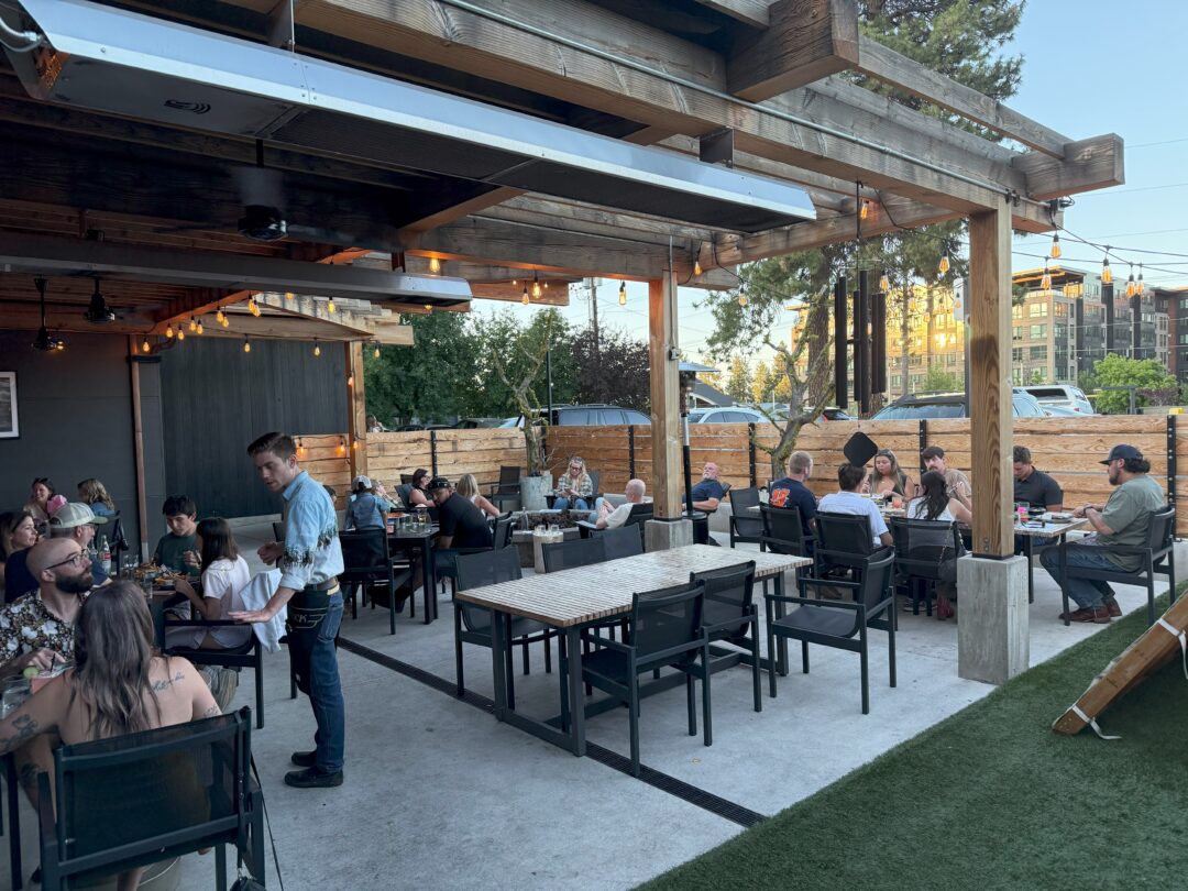 Outdoor restaurant patio with people dining at tables under a wooden pergola. String lights hang above. Some patrons are engaged in conversation, a waiter is serving food, and cars are visible beyond the wooden fence. Its early evening with clear skies.