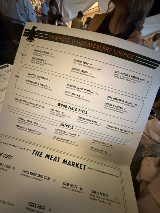 A person holds a large restaurant menu at a dimly lit table. The menu, titled Hawkeye & Huckleberry Lounge, lists starters, hot dishes, wood-fired pizza, entrees, and meats, with blurred diners visible in the background.