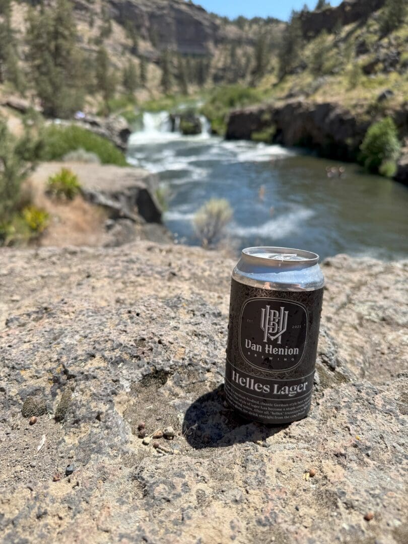 a Can of Dan Henion Helles Lager Sits on a Sunlit Rock at Steelhead Falls with a River Small Waterfall and Rocky Cliffs Framed by Green Trees in the Blurry Background Under a Clear Blue Skyan Inviting Scenic Outdoor Retreat