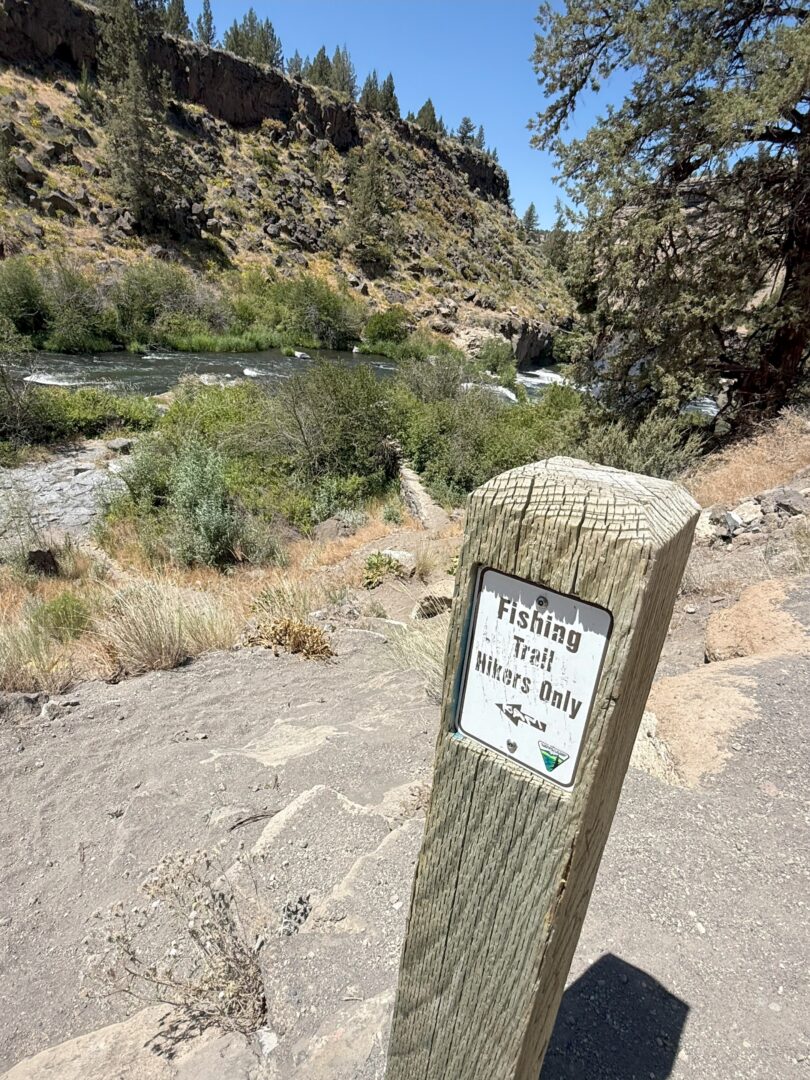 Hiking Trail to Steelhead Falls a Weathered Wooden Post with a Fishing Trail Hikers Only Sign Stands on a Dirt Path Overlooking Steelhead Falls Surrounded by Bushes Rocky Terrain and Tall Pine Trees Under a Bright Clear Blue Sky