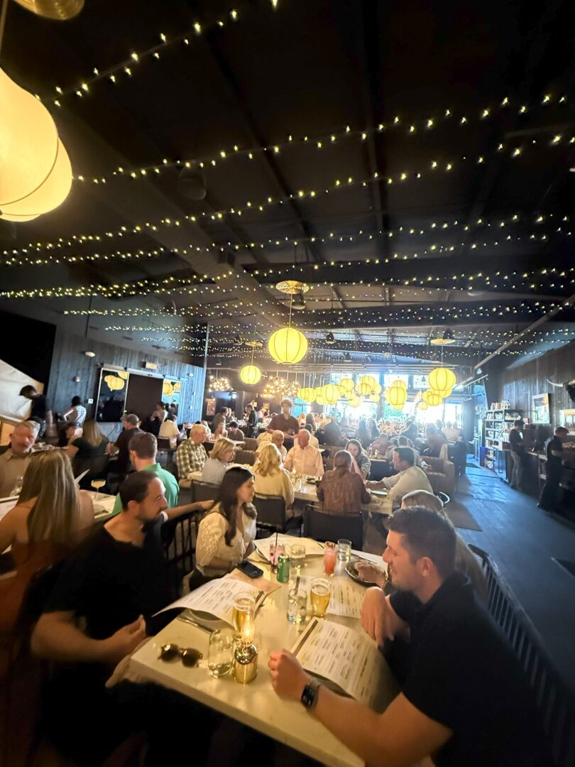A lively restaurant scene with many people dining at tables under warm hanging lights and string lights on the ceiling. The room is bustling with conversation, and menus and drinks are visible on the tables. The atmosphere appears cozy and inviting.