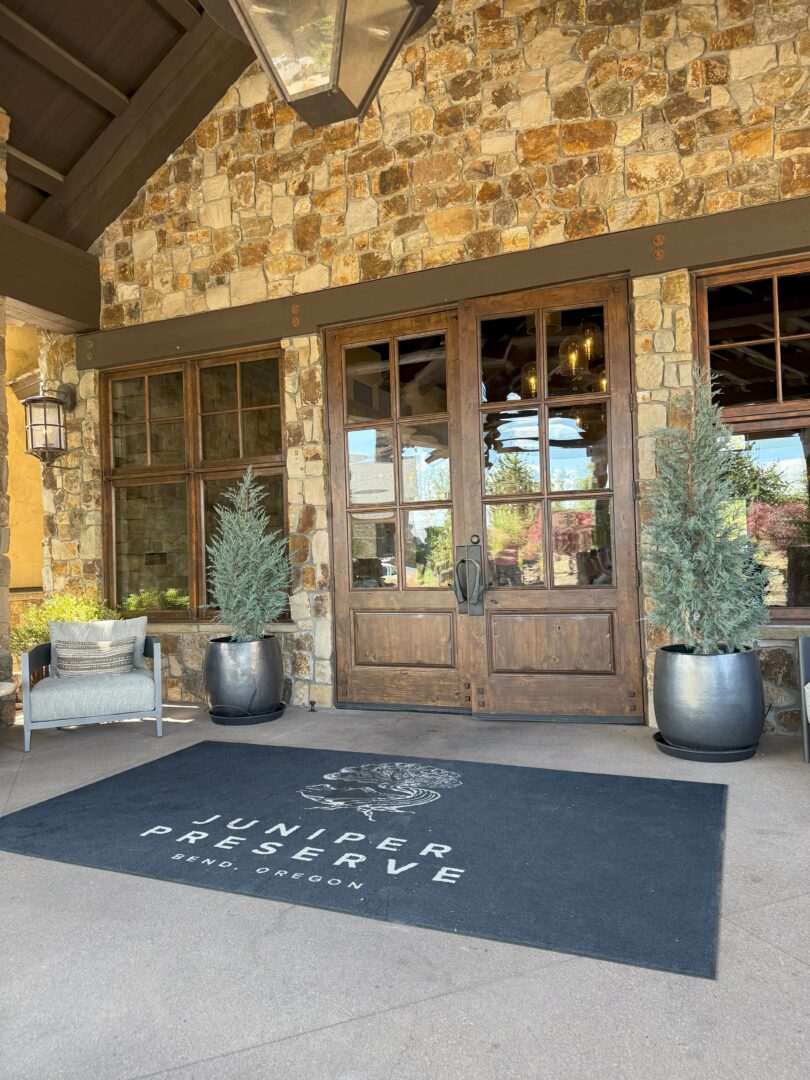 Juniper Reserve Bend Oregon a Stone Building Entrance with Double Wooden Doors Flanked by Large Potted Evergreens Welcomes Visitors to Pronghorn Golf Course a Black Mat in Front Reads Juniper Preserve Bend Oregon Nearby Sits a Grey Cushioned Chair and Lantern Style Wall Light
