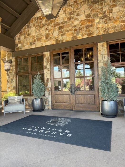 Juniper reserve bend oregon a Stone Building with Large Wooden Double Doors Flanked by Tall Potted Evergreen Trees Welcomes Guests Seeking Where to Stay in Bend Oregon a Black Rug Reads juniper Preserve Bend Oregon Nearby a Cushioned Chair and Side Table Sit Beneath a Wall Lantern