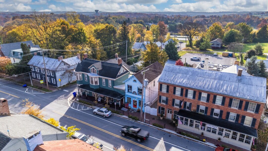 Main street new market md Aerial View of a Small Town Street Lined with Historic Buildings Some Brick and Some Painted Wood Surrounded by Autumn Trees Cars Are Parked Along the Street a Black Truck Passes By in the Distance Open Fields Stretch Toward Hotels in Frederick Md