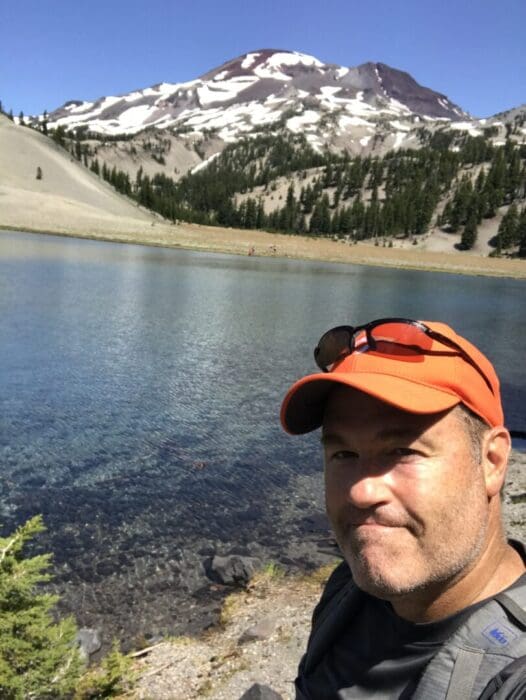 A man in an orange cap and sunglasses smiles near a clear blue alpine lake. Behind him, evergreen trees and a snow-dappled mountain rise under a bright blue sky, suggesting a sunny day in a scenic wilderness location.