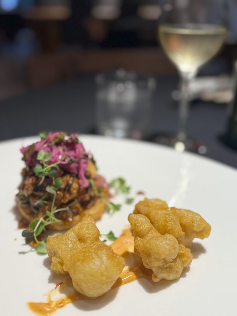 Mushroom tostada entree a White Plate at Market Table Bistro Holds Two Golden Crispy Fried Pieces Beside a Drizzle of Orange Sauce in the Background Shredded Meat is Topped with Purple Slaw and Microgreens While Wine and Water Glasses Sit Blurred in the Distance