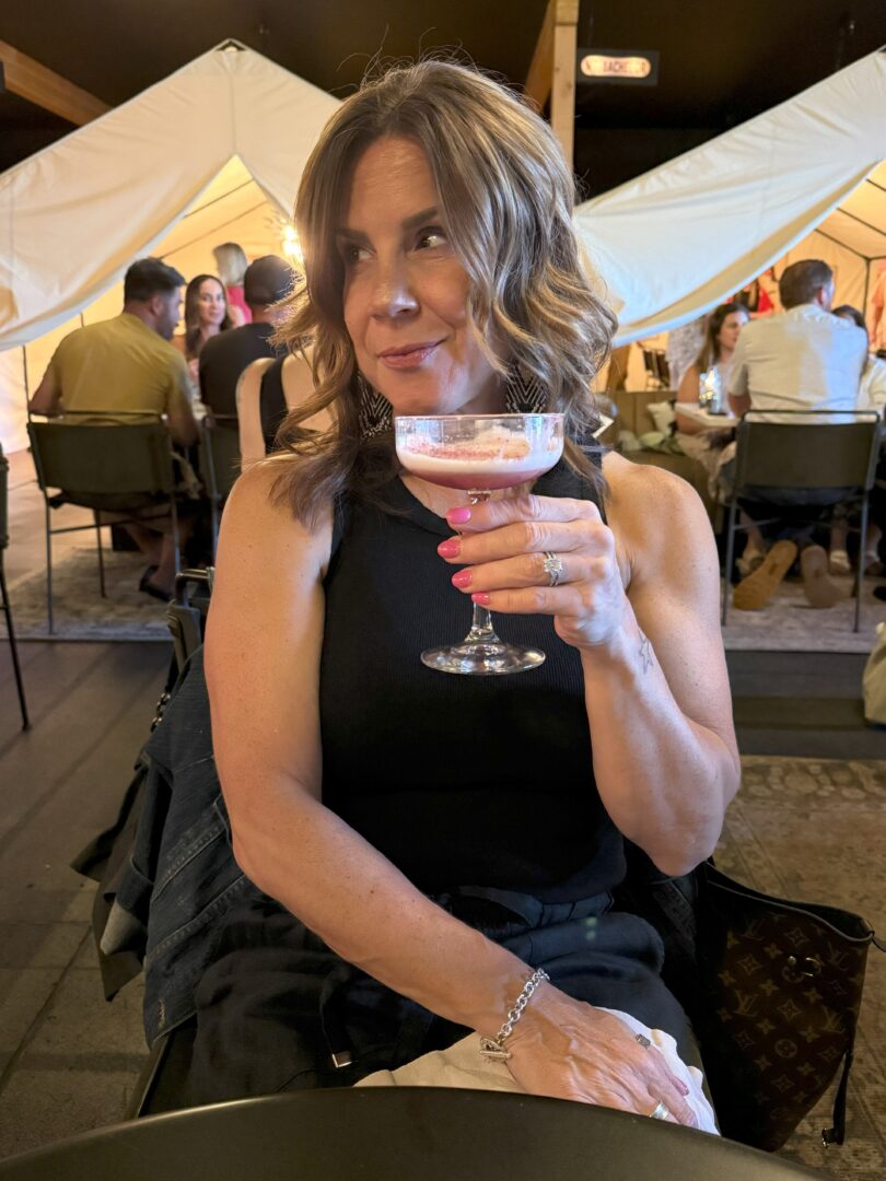 A woman with wavy brown hair in a black sleeveless top sits at a table, holding a pink cocktail in a coupe glass, looking thoughtfully to her left. Behind her, people sit at tables in a warmly lit tented setting, suggesting a social gathering.