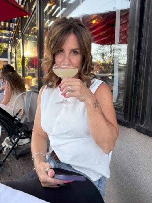 a Woman with Wavy Brown Hair and a Sleeveless White Top Sits Outdoors at a Restaurant Holding a Cocktail Glass Up to Her Mouthperhaps Enjoying the Vibrant Scene at One of the Bars in Bend Or a Stroller is Visible in the Background