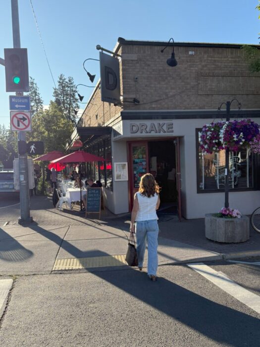 Nicole Drake Restaurant Bend or a Woman in a White Top and Light Blue Jeans Walks Toward the Entrance of Drake Restaurant in Downtown Bend Oregon on a Sunny Day Outdoor Diners Sit Under Red Umbrellas As She Explores Airbnb Bend Oregon Nearby Hanging Flower Baskets and a Green Traffic Light Are Visible