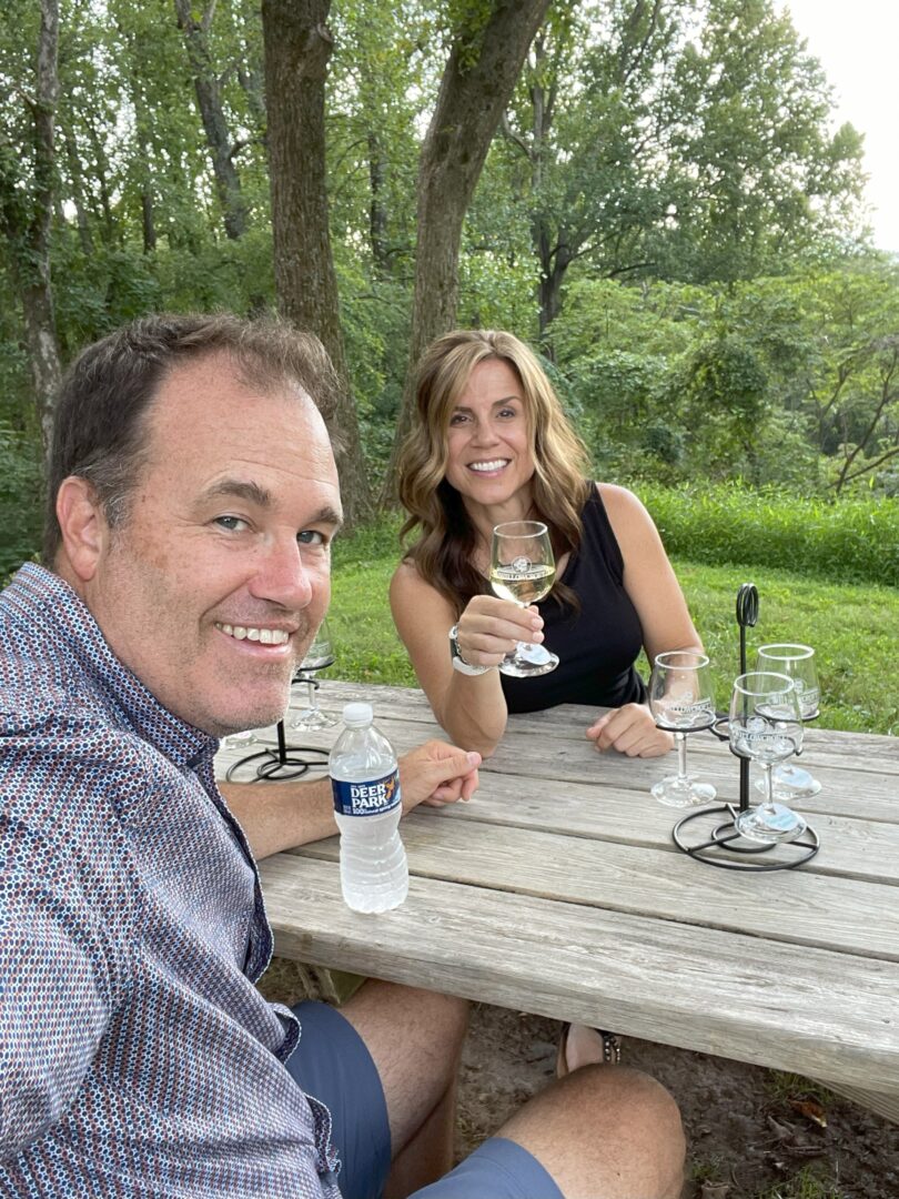 Nicole glenn wine tasting loudoun county a Man and Woman Sit at a Wooden Picnic Table Outdoors at Market Table Bistro Smiling at the Camera the Woman Holds a Wine Glass with More Empty Glasses on the Table the Man Holds a Water Bottle Amid Lush Green Trees and Grass on a Sunny Day
