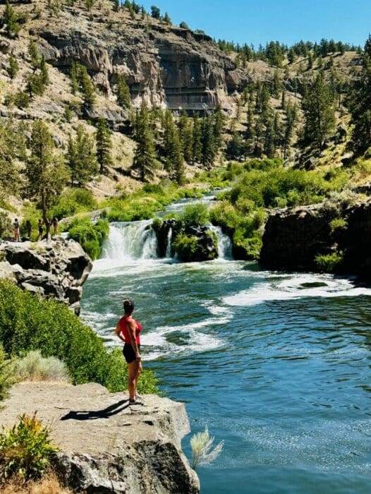 A person in a red shirt and black shorts stands on a rocky ledge by Steelhead Falls, gazing at a small waterfall. Green trees and shrubs surround the scene, with steep, rocky hills under a clear blue sky in the background.