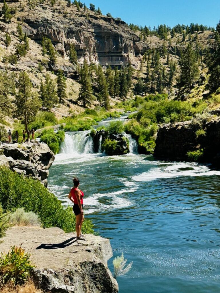 Nicole Steelhead Falls a Person in a Red Shirt and Black Shorts Stands on a Rocky Ledge by Steelhead Falls Gazing at a Small Waterfall Green Trees and Shrubs Surround the Scene with Steep Rocky Hills Under a Clear Blue Sky in the Background