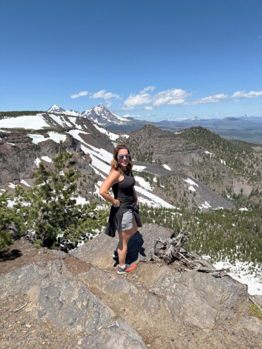 Nicole Tam Mcarthur Rim Trail Summit a Woman in Athletic Clothing and Sunglasses Stands Smiling on a Rocky Mountain Ledge Behind Her Are Snowy Peaks Green Forests and a Clear Blue Skycapturing the Adventure of Hiking Near an Airbnb in Bend Oregons Stunning Wilderness