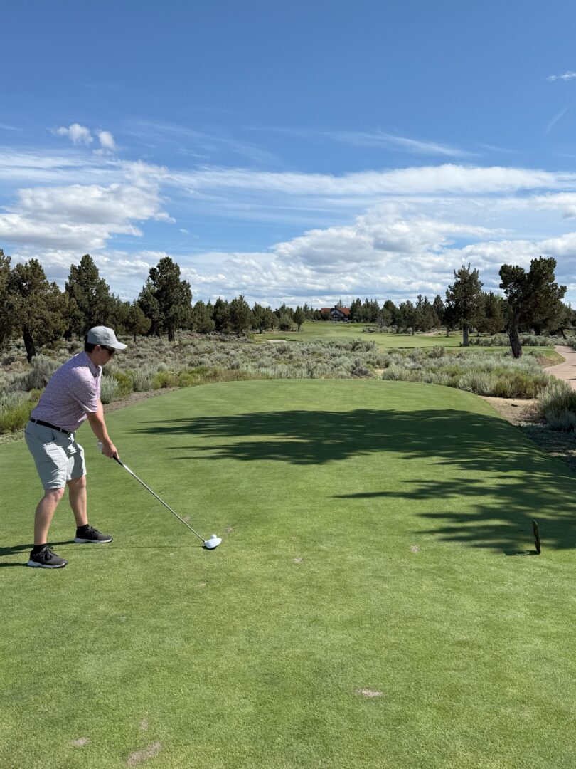 Pronghorn Golf Club Bend Oregon a Person Wearing a Cap Light Shirt and Shorts Stands on a Tee at Pronghorn Golf Course Preparing to Swing a Golf Club the Green Fairway Stretches Ahead Surrounded by Trees and Shrubs Under a Blue Sky with Scattered Clouds