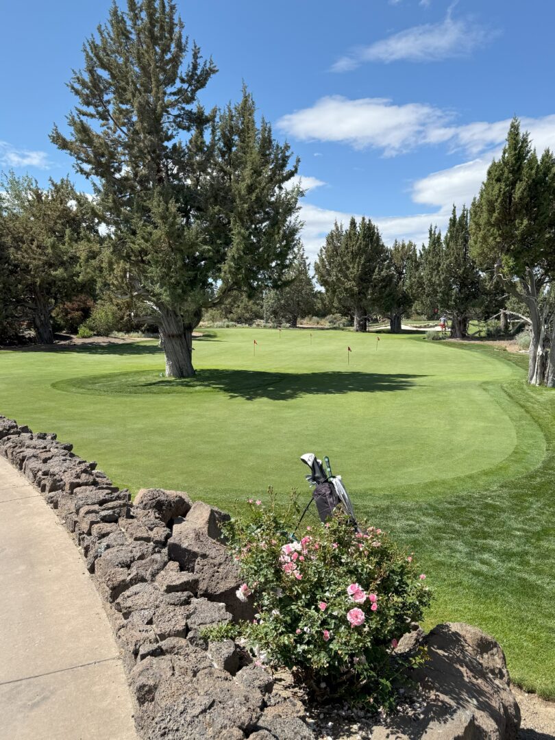 Putting Green Pronghorn Golf Course a Golf Green at Pronghorn Golf Course is Bordered by Trees and a Stone Wall with a Golf Bag Leaning Against It Pink Flowers Bloom in the Foreground While Sunlight Filters Through Partly Cloudy Skies Onto the Well Kept Grass and Flags