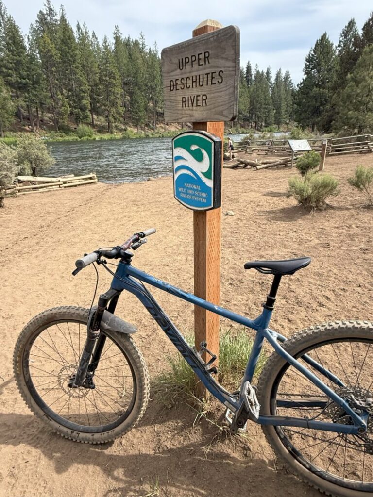 A blue mountain bike is parked on a dirt path by a wooden sign reading “Upper Deschutes River” beside the flowing river, with pine trees and fences nearby. The Rimrock Trail runs under a partly cloudy sky.